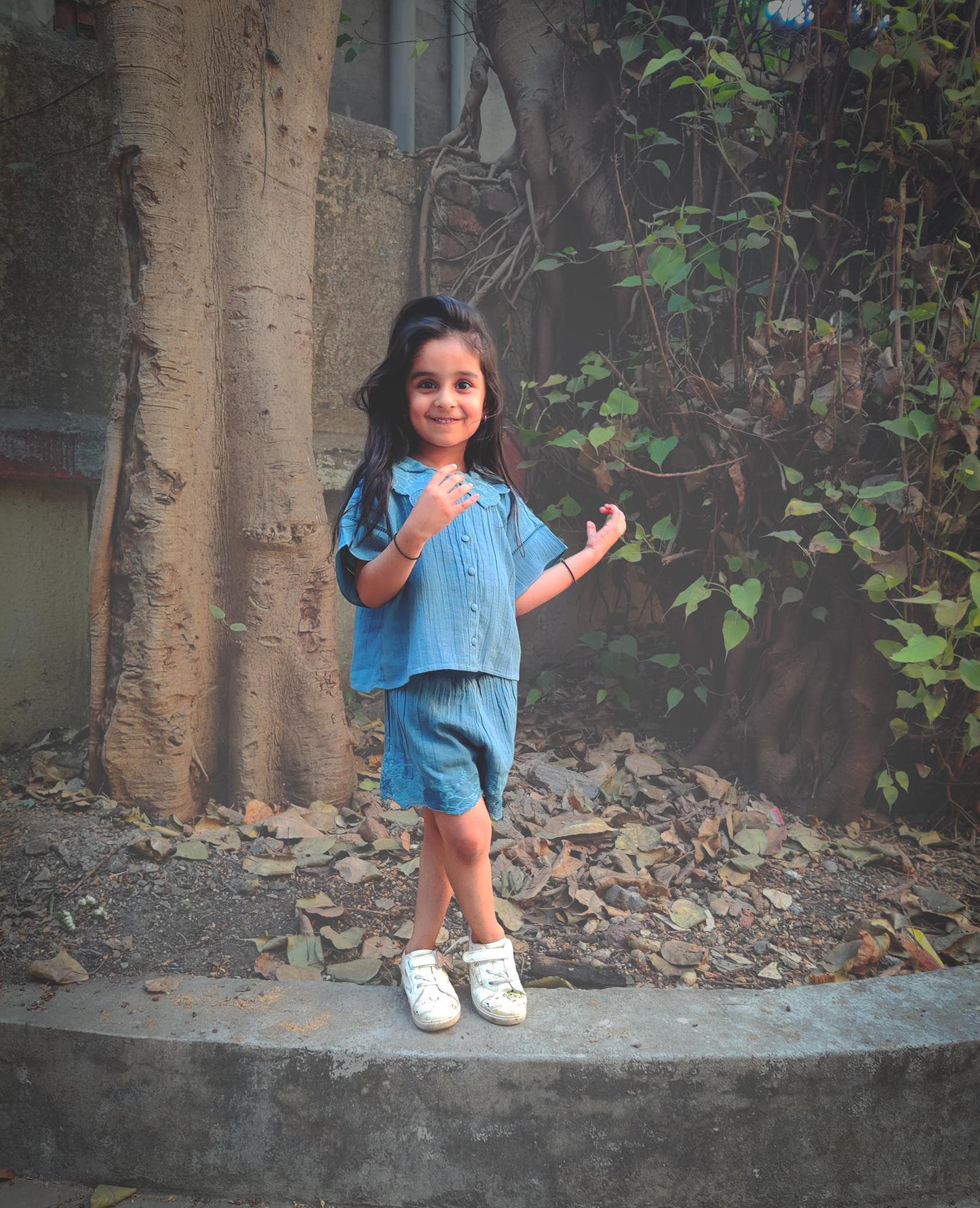 Happy child wearing a blue collared summer outfit from The Kefi House and white shoes, standing outdoors.