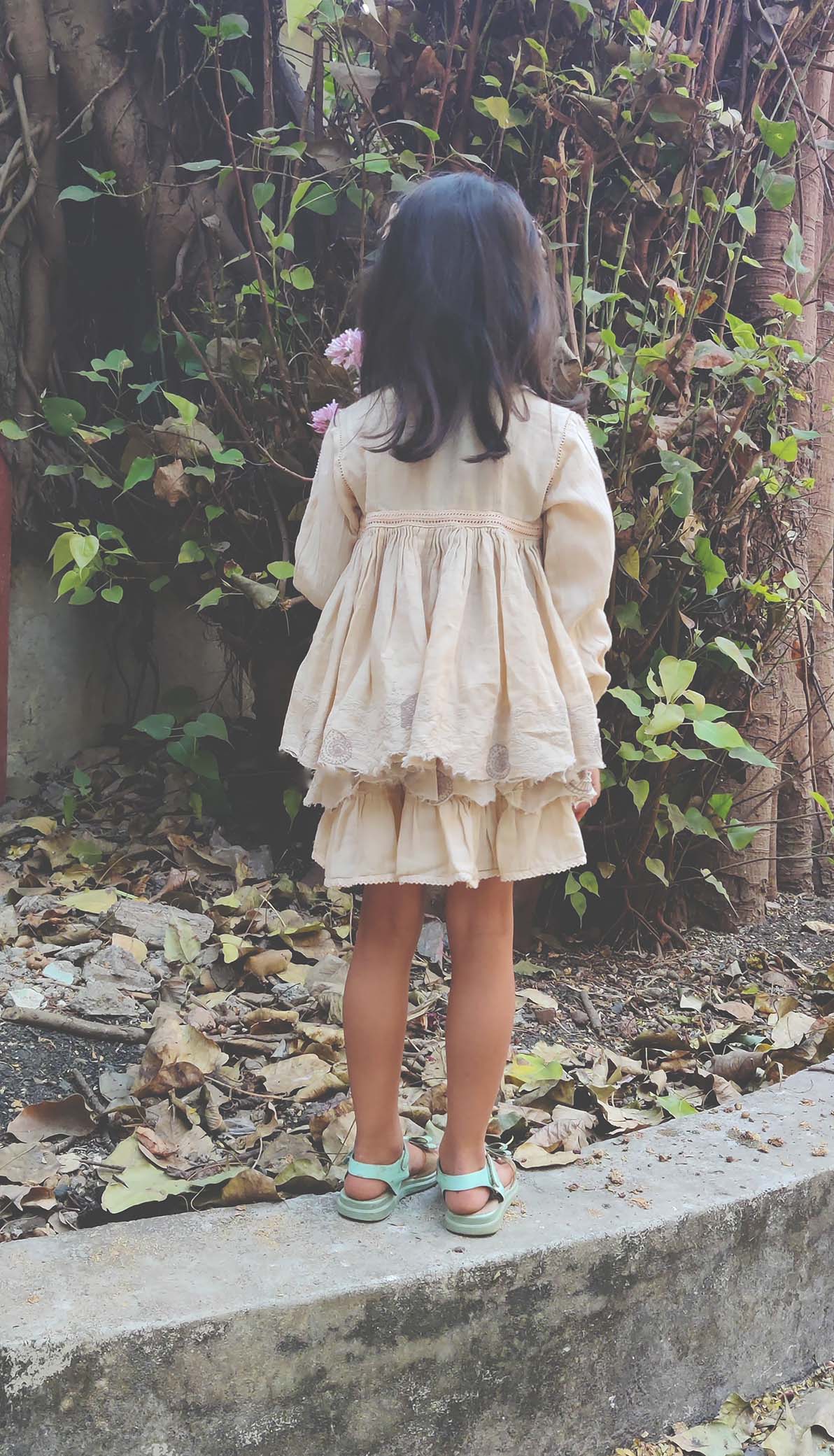 Back view of a young girl wearing a cream-coloured layered cotton dress with lace detailing at the hem and waist. The dress features long sleeves, a gathered skirt, and a relaxed, flowy fit. She is standing outdoors against a background of green foliage and dry leaves.