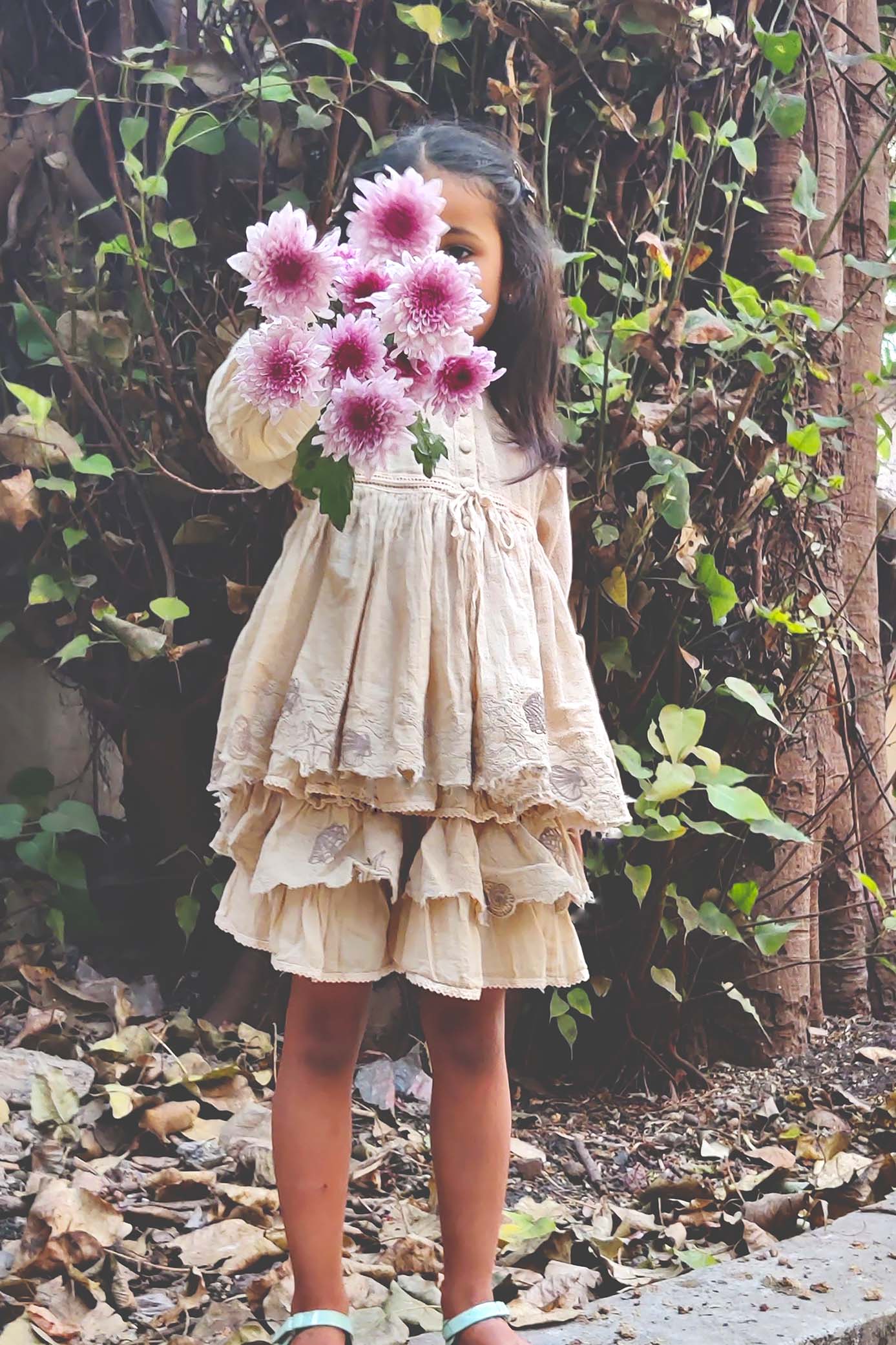 Front view of a young girl wearing a layered off-white cotton dress with delicate lace detailing and gathered tiers. She is standing outdoors on dry leaves, holding a bouquet of pink flowers in front of her face, surrounded by green foliage in the background.