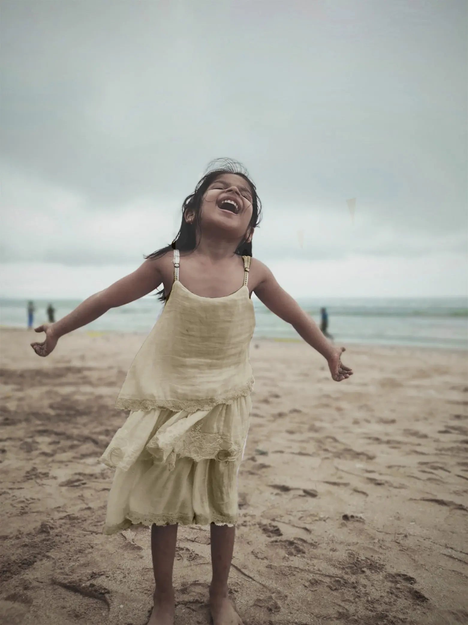 A happy girl in a natural-toned dress playing on the beach with the waves behind her