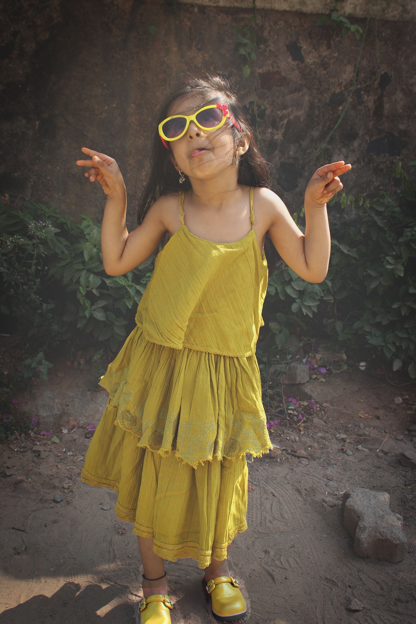 Young girl wearing golden yellow layered cotton dress with lace hem and yellow sunglasses, posing playfully outdoors