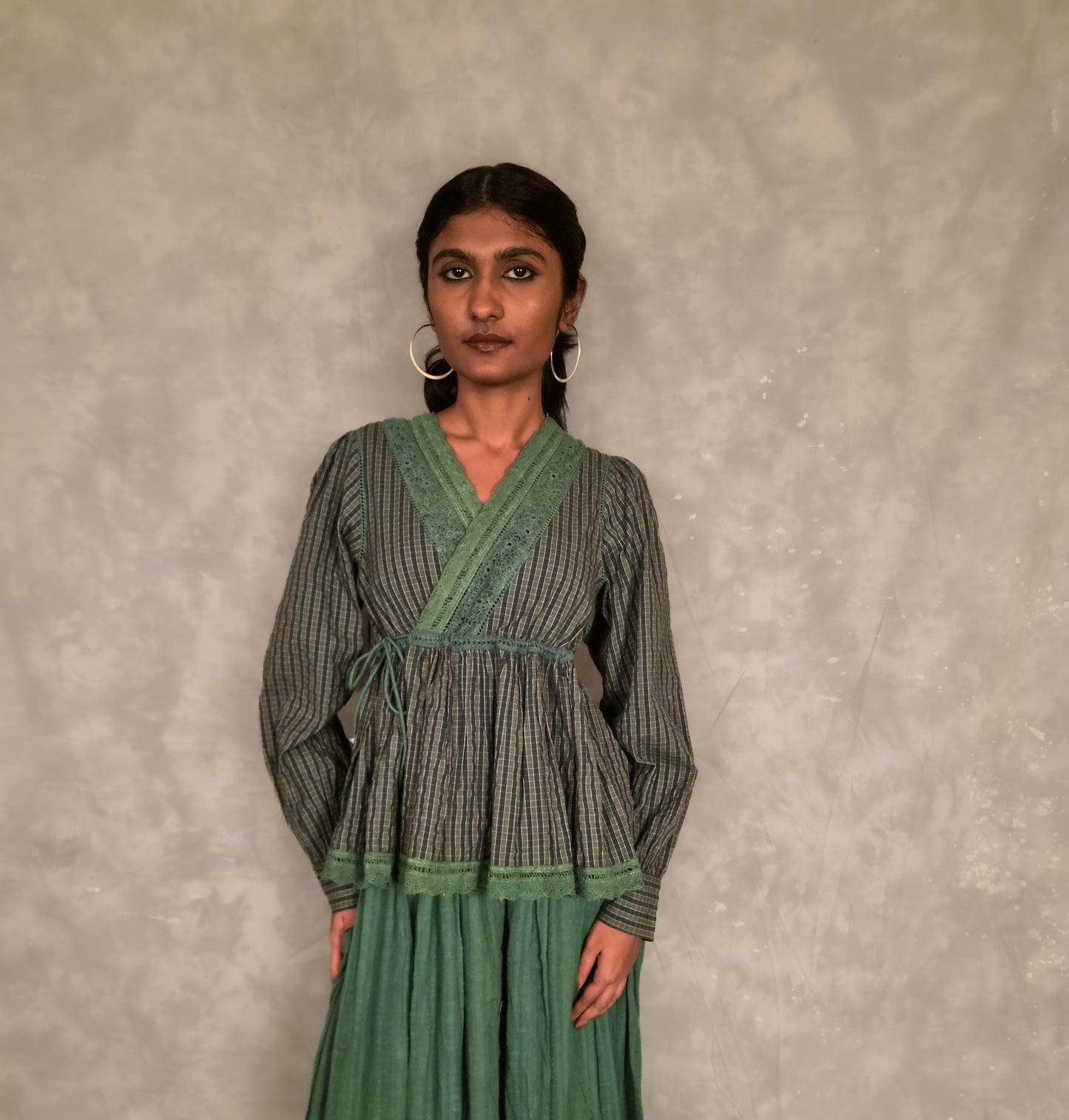 A woman modeling a long-sleeved green and gray checkvwrap-style top with a matching pleated green skirt, standing against a textured gray wall.