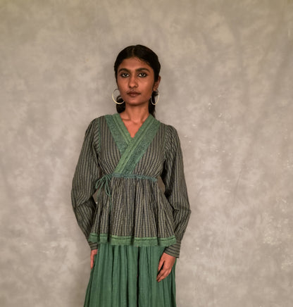 A woman modeling a long-sleeved green and gray checkvwrap-style top with a matching pleated green skirt, standing against a textured gray wall.